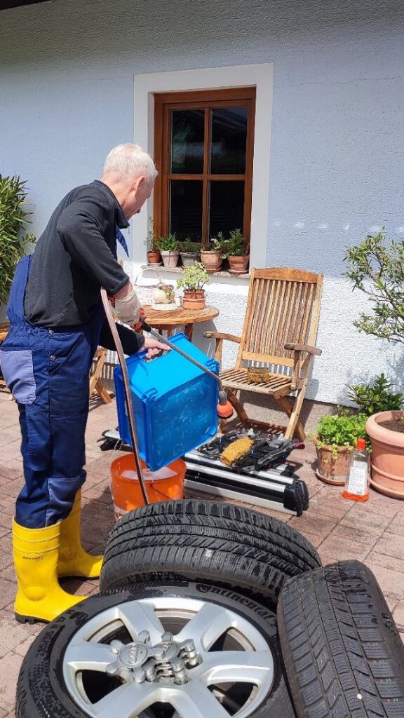 ein Mann in Latzhose und Gummistiefel hantiert mit Putzkübel und Gartenschlauch. Er wäscht Reifen, Dachträger und eine Plastikkiste