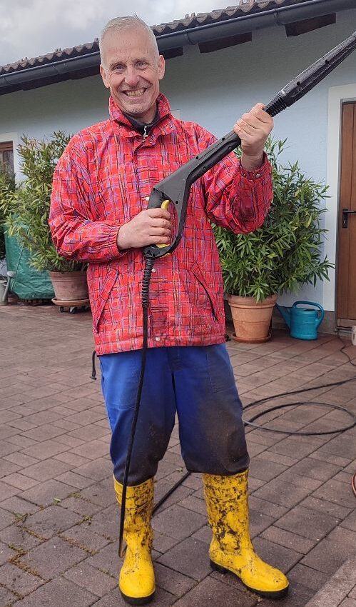 Ein Mann mit einer Hochdruckreinigerpistole posiert fürs Foto in nasser, schmutziger Hose und und schmutzigen Gummistiefeln 