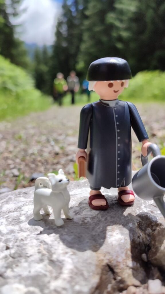 Im Vordergrund steht eine Playmobilfigur, die Sebastian Kneipp im Talar darstellt mit seinem Hund, einem Spitz auf einem Stein. Im unscharfen Hintergrund sieht man 3 Personen schemenhaft auf einem Kiesweg gehen