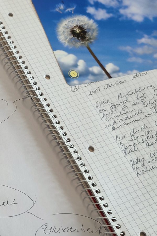 ein Spiralblock mit handgeschriebenen Schriftfragmenten und daraufliebendem Foto mit einer Pusteblume 