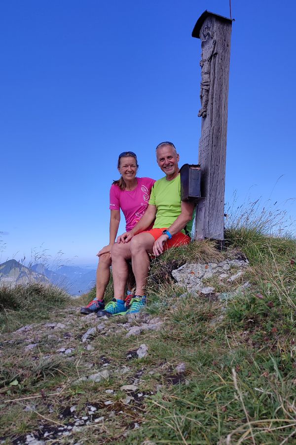 eine lächelnde Frau und ein lächelnder Mann sitzen neben einem Gipfelkreuz aus Holz auf einem Gipfel - im Hintergrund sieht man Berge
